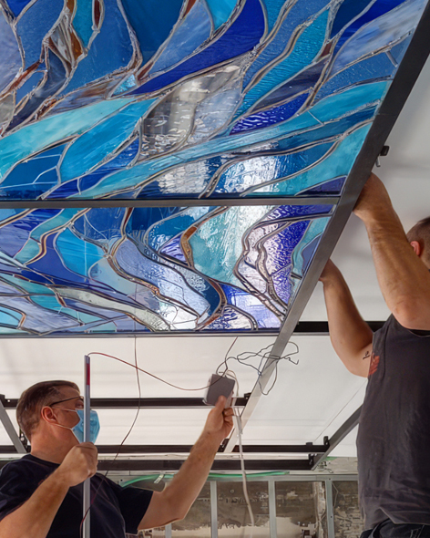 שני אנשים מתקינים לוח תקרה גדול מזכוכית צבעונית עם דוגמאות כחולות מסולסלות. אדם אחד מחזיק מקל מדידה, בעוד האחר מכוון את הלוח. שניהם עובדים בזהירות, כשכלי העבודה והחוטים נראים לעין.