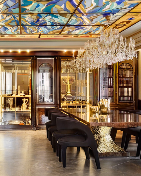 חדר אוכל מפואר עם שולחן ארוך ומלוטש, כיסאות שחורים, נברשת קריסטל, תקרת ויטראז' עם דוגמאות מופשטות וארונות זכוכית המציגים פריטי נוי אלגנטיים. תאורה חמימה מדגישה את האווירה המפוארת.