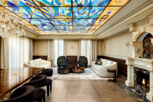 סלון אלגנטי עם שתי ספות לבנות, כורסאות שחורות, אח משיש ותקרת ויטראז' צבעונית עם דוגמאות בכחול וצהוב. חלונות גדולים עם וילונות שקופים ונברשת משלימים את העיצוב המפואר.