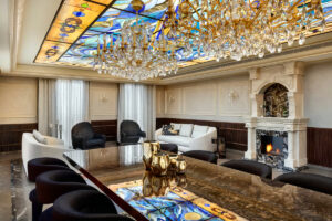 סלון מפואר כולל תקרת ויטראז', נברשות מפוארות, ספות וכורסאות אלגנטיות, אח משיש עם אש בוערת ושולחן גדול ומלוטש עם קישוטים מזהב. אור טבעי נכנס מבעד לווילונות שקופים.