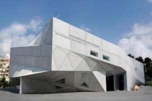 בניין מודרני וזויתי עם לוחות בטון גיאומטריים וחלונות צרים, ובו הכניסה למוזיאון תל אביב לאמנות מתחת לקטע בולט, על רקע שמיים כחולים עם כמה עננים.