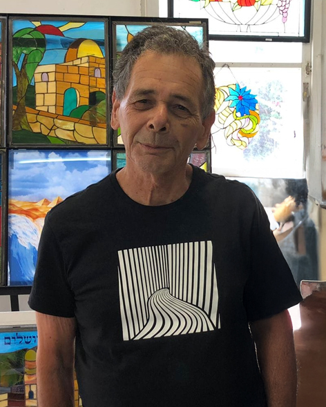 גבר עם שיער אפור קצר עומד בתוך הבית, לבוש בחולצה שחורה עם הדפס לבן בגלים. מאחוריו מוצגת יצירת אמנות מזכוכית צבעונית עם דוגמאות וצבעים ססגוניים. אור זורח מבעד לחלון ברקע.