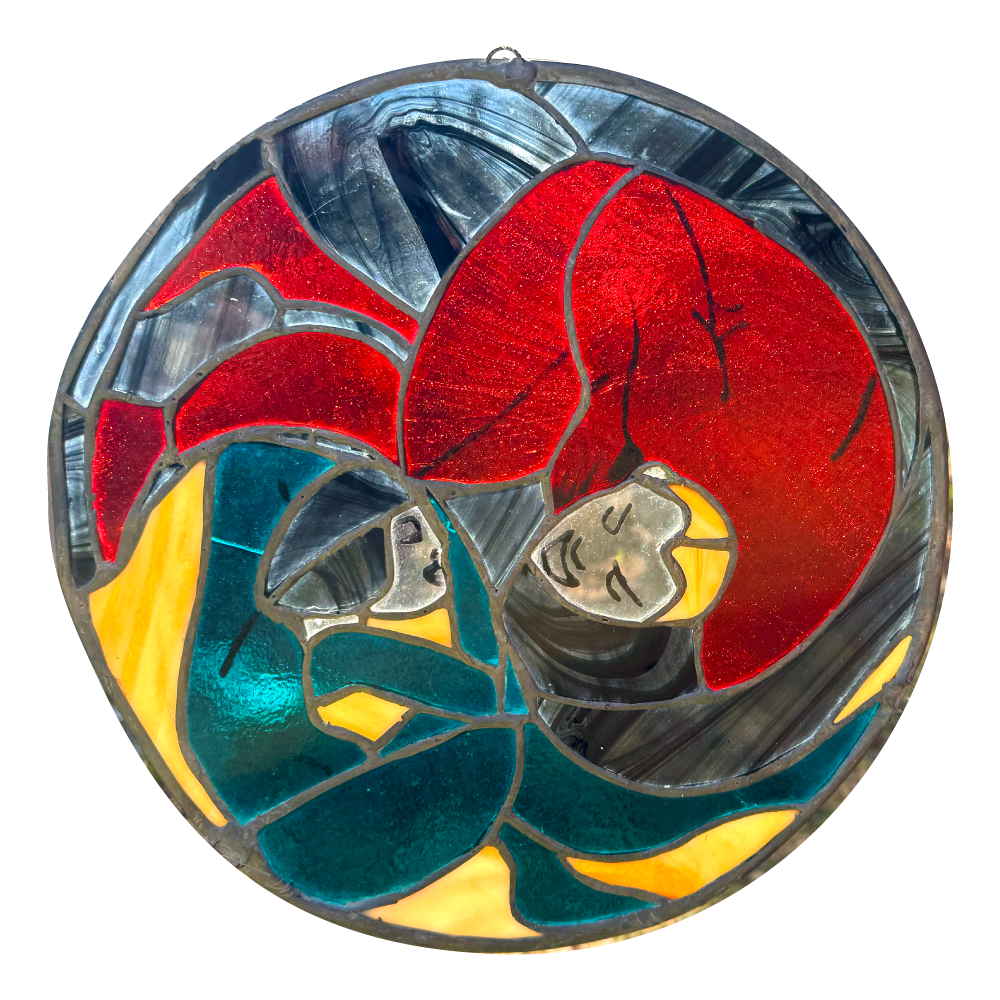 יצירת אמנות עגולה מזכוכית צבעונית המציגה שתי דמויות אנושיות מופשטות ושזורות זו בזו, האחת עם שיער אדום בוהק והשנייה עם שיער בצבע טורקיז, על רקע של חתיכות זכוכית כחולות, צהובות ואפורות.