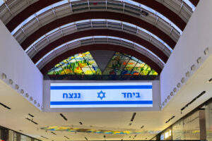 מסך דיגיטלי גדול בקניון מציג את דגל ישראל וטקסט בעברית. מעליו, חלון ויטראז' צבעוני מעוטר בעיצובים שונים. תקרת הקניון מקושתת ומוארת בתאורה בהירה.