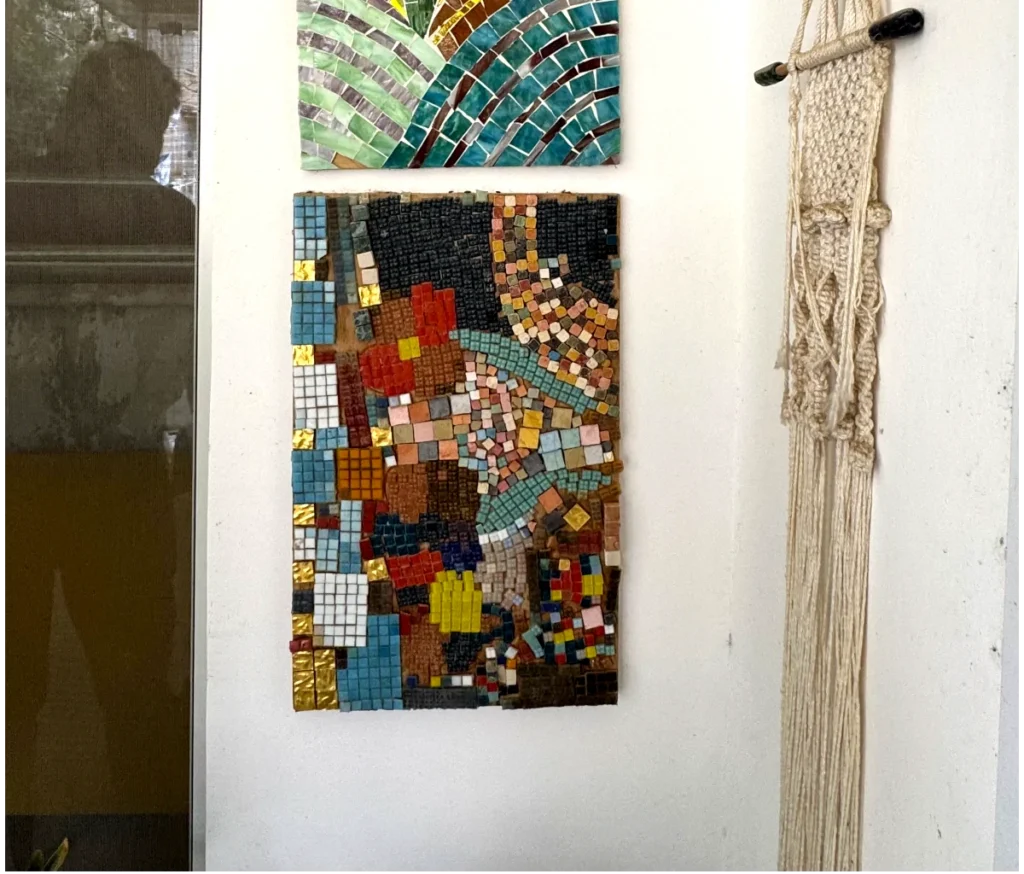 היצירה "מפגש צבעים" — פסיפס צבעוני של אריחים קטנים ומרובעים — מוצגת על קיר לבן לצד יצירת מקרמה ארוגה ומתחת לפסיפס ירוק-כחול נוסף, כשמשמאל נראית השתקפות קלושה של חלון.