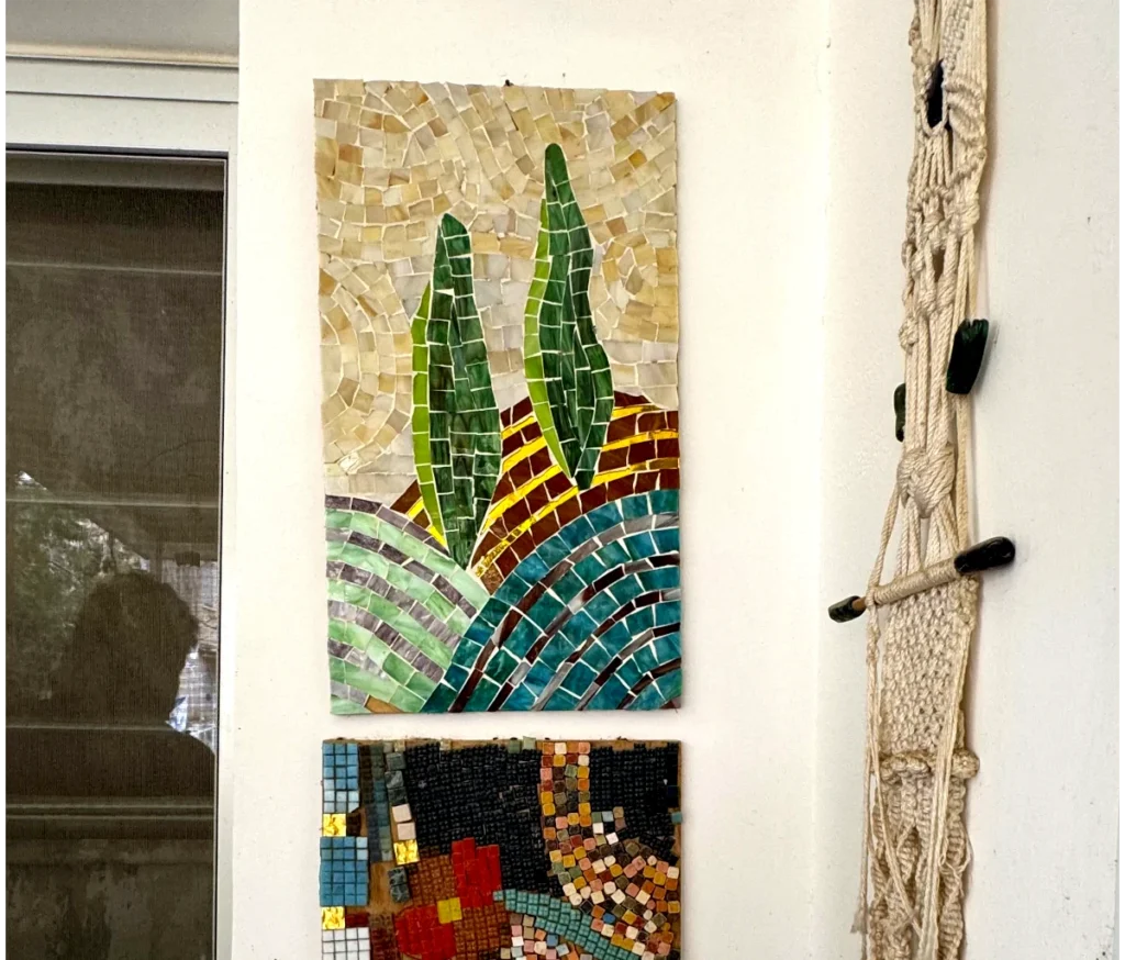 היצירה "ברושים וגבעות בפסיפס אומנותי" — פסיפס צבעוני של שני עצים ירוקים על גבעות — תלויה על קיר לבן לצד מקרמה ופסיפס מופשט, כשמשמאל נראית השתקפות של חלון.