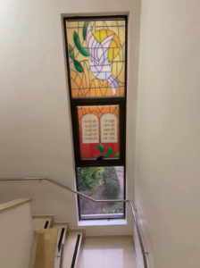 חלון ויטראז' מעל גרם מדרגות, ובו יונה לבנה ועלים ירוקים בחלקו העליון וטקסט בעברית בחלקו התחתון, כשדרך לוח הזכוכית השקוף התחתון נראית נוף של צמחייה.