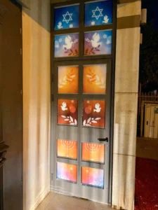 דלת עם לוחות צבעוניים המעוטרים במגני דוד, יונים לבנות, ענפי זית, רימונים ומנורות, המוארת בלילה ומוקפת בקיר אבן.