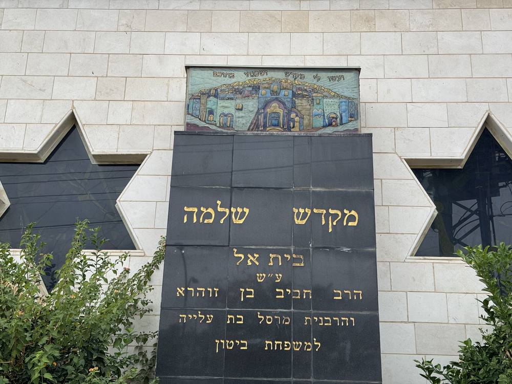 בקיר האבן יש שני חלונות גדולים בצורת כוכב ולוחית עם כיתוב בעברית. מעל הלוחית יש פסיפס אריחים המציג מבנה, שגם עליו כתוב בעברית. שיחים ירוקים צומחים מול הלוחית.