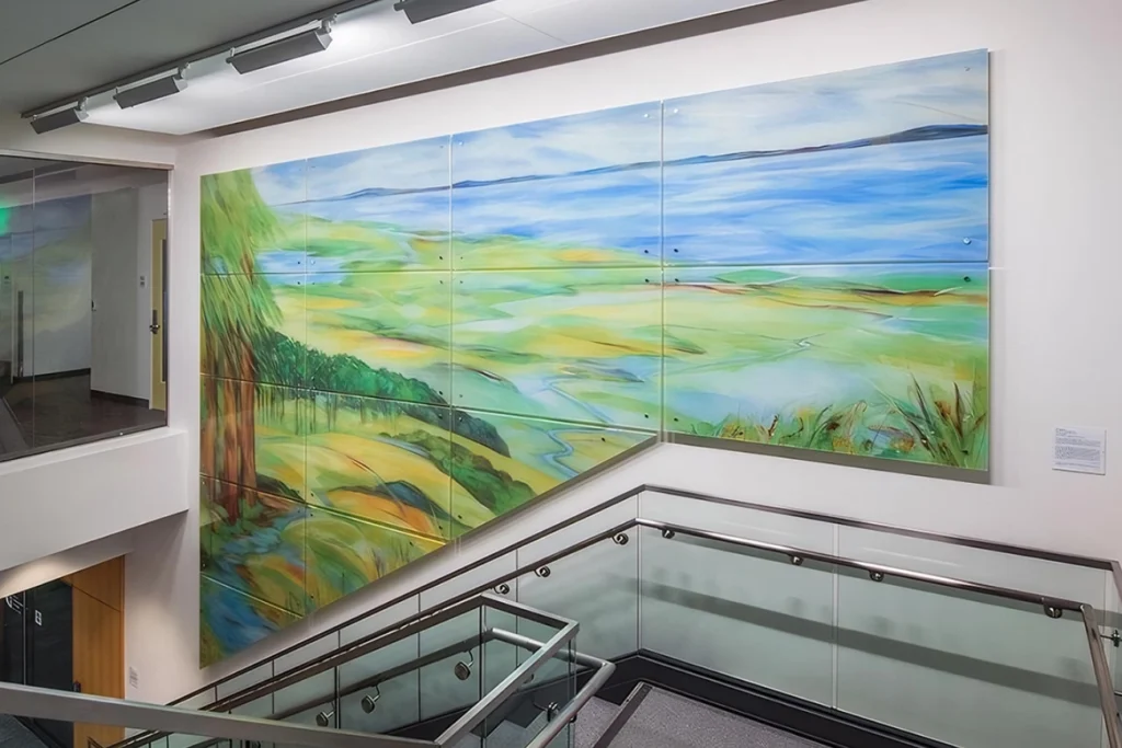 על הקיר מעל גרם מדרגות מודרני עם מעקות זכוכית ומתכת מתנוססת ציור קיר נופי גדול וצבעוני, המציג שדות, עצים ושמיים כחולים.