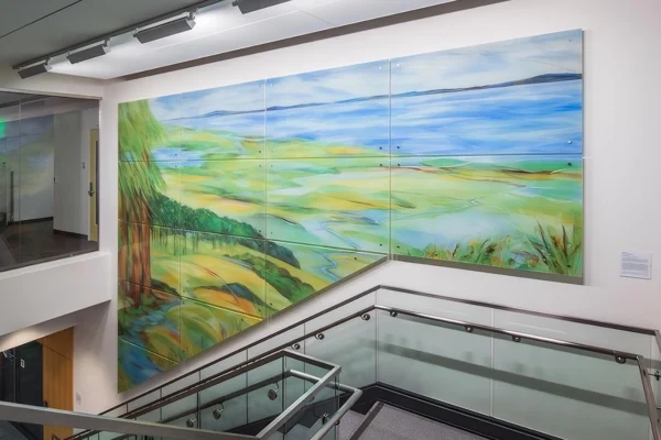 על הקיר מעל גרם מדרגות מודרני עם מעקות זכוכית ומתכת מתנוססת ציור קיר נופי גדול וצבעוני, המציג שדות, עצים ושמיים כחולים.
