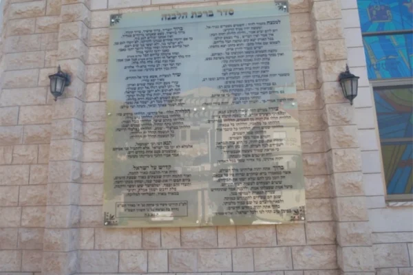 לוח זכוכית גדול עם כיתוב בעברית מותקן על קיר אבן בין שני פנסים שחורים. על הזכוכית נראים השתקפויות של מבנים ושל השמיים. מימין נראית חלקית יצירת אמנות מזכוכית צבעונית.